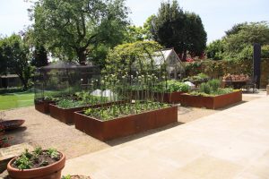 six corten steel raised planters sitting on a gravel garden with green plants growing inside and an archway in the middle