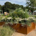 Large rusty orange corten steel planter with green plants growing inside at a garden show