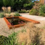 A corten steel pond set in the ground with a corten water rill at both ends flowing water inside and stones around the outside in a garden