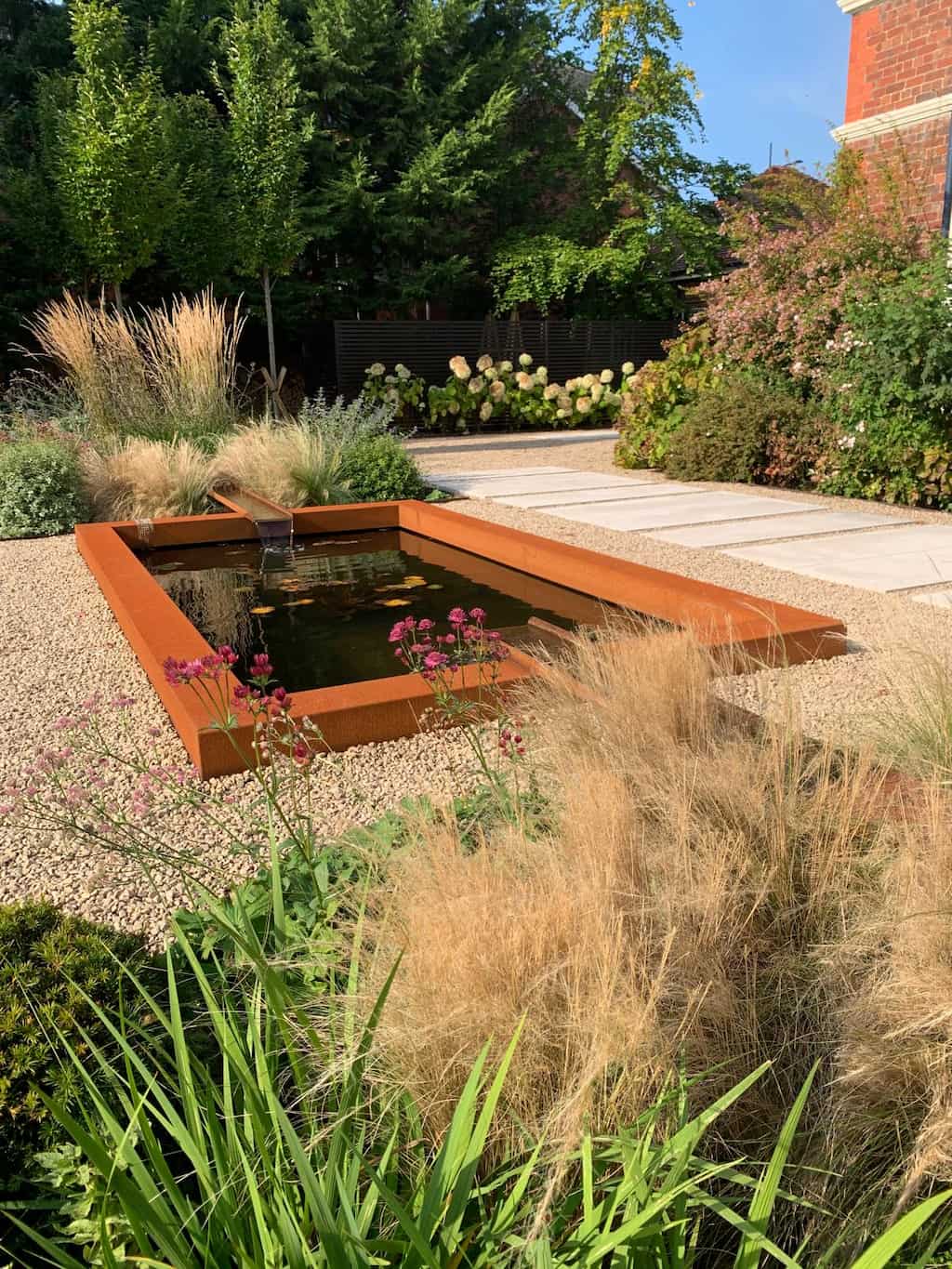 A corten steel pond set in the ground with a corten water rill at both ends flowing water inside and stones around the outside in a garden