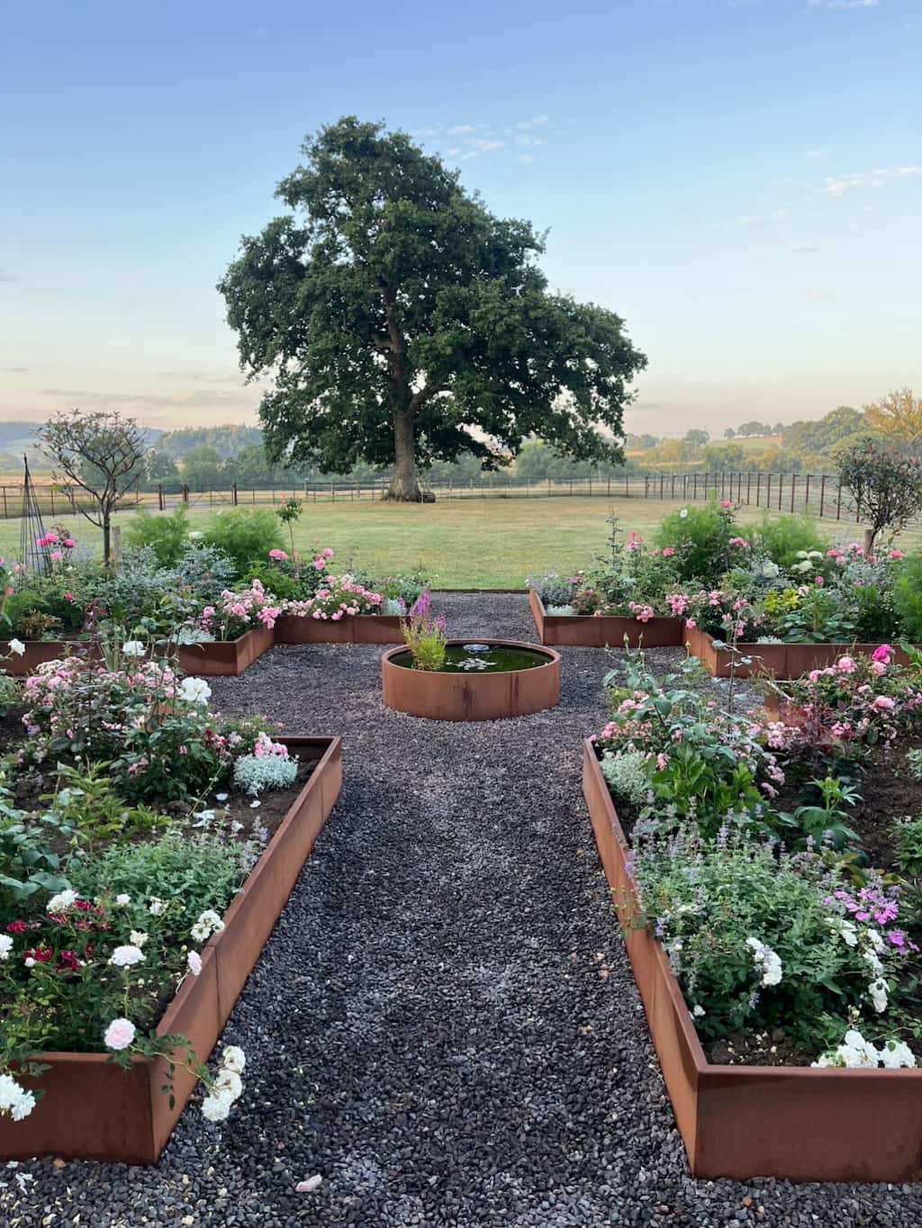 Corten steel raised beds with Circular water feature Corten steel raised beds with plants inside and a corten round water pond in the middle with a large tree and garden in the background
