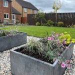 Galvanised steel planter with plants inside sitting on top of stones with a brown fence and grass behind