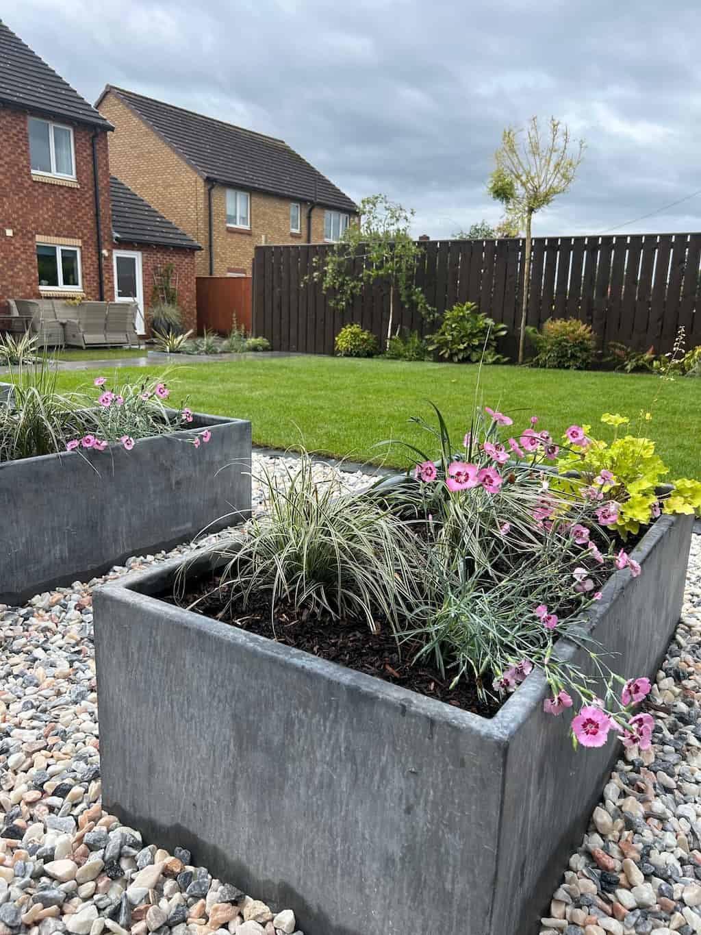 Galvanised steel planter with plants inside sitting on top of stones with a brown fence and grass behind