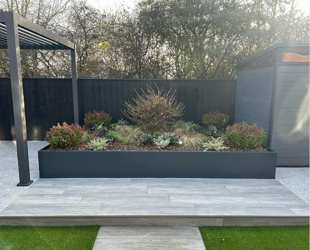 Powder coated trough planter set inside decking Anthracite grey powder coated steel planter with plants growing inside set into raised decking with a black fence and trees in the background