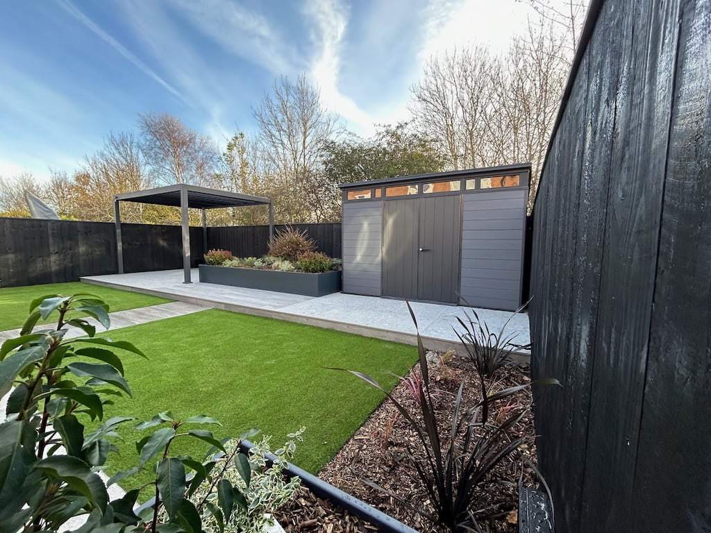 Powder coated trough planter with pergola Anthracite grey steel planter with plants growing inside including a pergola on the left and a garden shed on the right in a garden setting