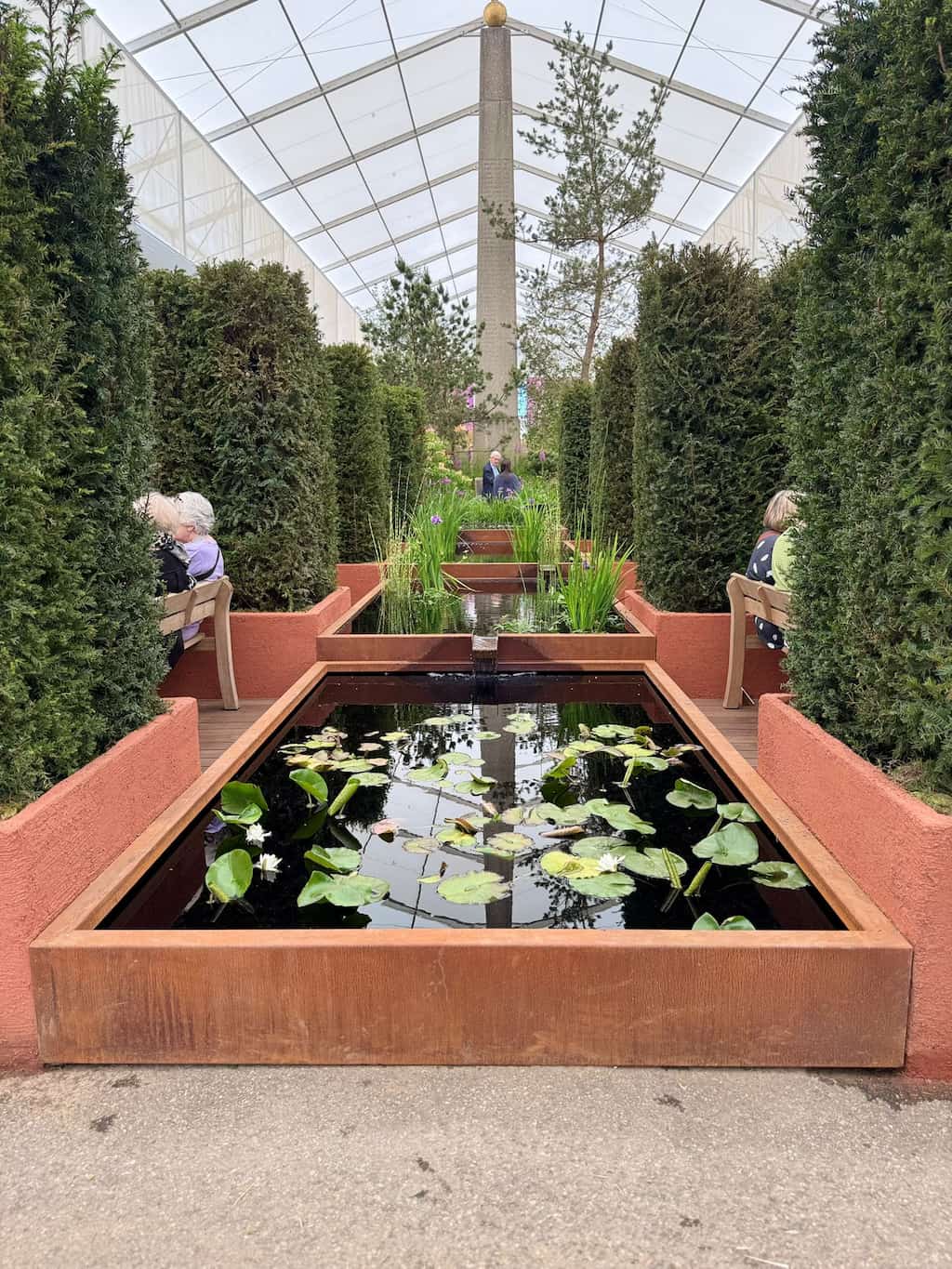 RHS chelsea 2025 corten water feature 2 3 Corten steel water ponds in a row with green plants on both sides and people sat on benches at the RHS Chelsea Flower show 2025
