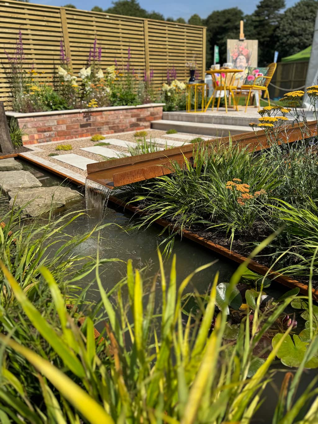 RHS show garden corten steel rill 2 Corten steel rill with water flowing into a corten steel pond with green plantes on either side and a table and chairs in the background