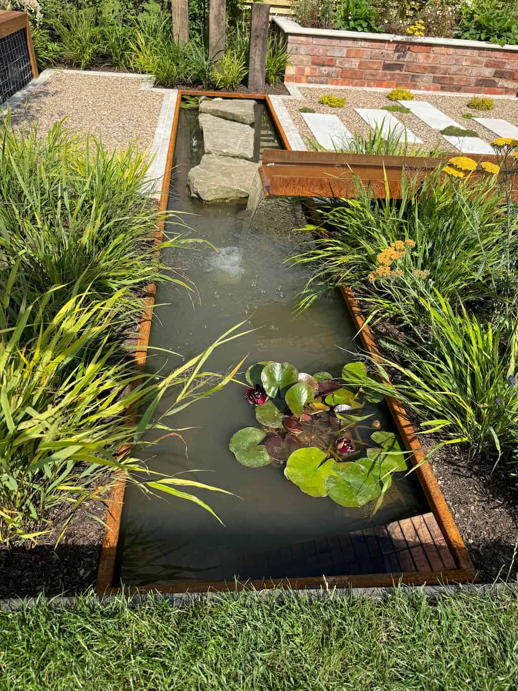RHS show garden corten steel water feature 3 A rusty steel water rill with flowing water into a water pond set in the ground with green plants at both side